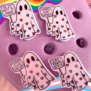 May include: Four ghost-shaped charms with heart accents and various text signs. The signs read: "BI IS NOT A PHASE", "GHOULS LOVE GHOULS", "TRANS LIVES MATTER", and "GAY RIGHTS BITCH". The charms are on a lavender surface with rainbow and transgender flag accents.