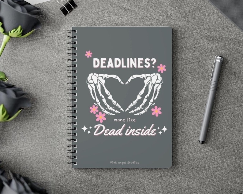 May include: Gray spiral bound notebook with a black skeleton hand design forming a heart shape. The text reads "Deadlines? More like Dead inside." with pink flowers and glitter accents. The bottom of the notebook says "Pink Angel Studios."