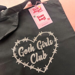 Puede incluir: Bolsa de tela negra con un dise&ntilde;o de brillo plateado en forma de coraz&oacute;n rodeado de alambre de p&uacute;as. El texto "Goth Girls Club" est&aacute; dentro del coraz&oacute;n. Una etiqueta rosa con el texto "Pink Angel Studios Tote Bag" est&aacute; unida a la bolsa.