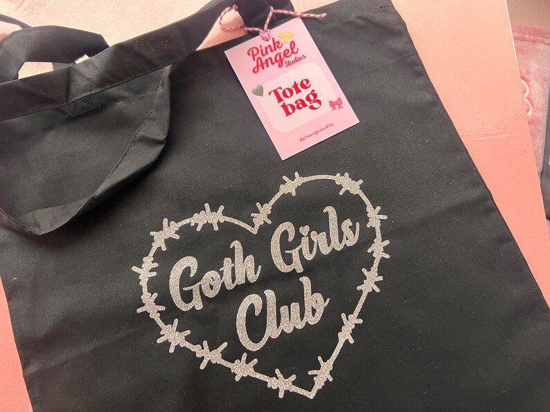 Puede incluir: Bolsa de tela negra con un dise&ntilde;o de brillo plateado en forma de coraz&oacute;n rodeado de alambre de p&uacute;as. El texto "Goth Girls Club" est&aacute; dentro del coraz&oacute;n. Una etiqueta rosa con el texto "Pink Angel Studios Tote Bag" est&aacute; unida a la bolsa.