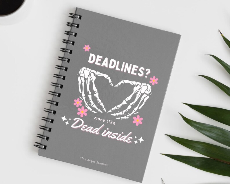 May include: Gray spiral bound notebook with a white skeleton hand design forming a heart shape. The text "Deadlines? more like Dead inside" is printed in pink on the front cover.
