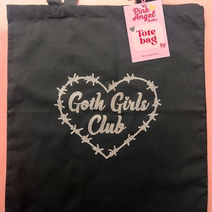 Puede incluir: Bolsa de tela negra con un dise&ntilde;o de coraz&oacute;n con alambre de p&uacute;as a su alrededor y el texto "Goth Girls Club" en purpurina plateada. La bolsa tiene una etiqueta que dice "Pink Angel Studios Tote Bag".