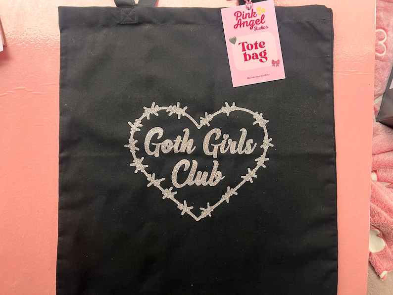 Puede incluir: Bolsa de tela negra con un dise&ntilde;o de coraz&oacute;n con alambre de p&uacute;as a su alrededor y el texto "Goth Girls Club" en purpurina plateada. La bolsa tiene una etiqueta que dice "Pink Angel Studios Tote Bag".