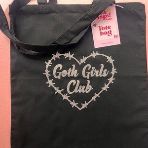 Puede incluir: Bolsa de tela negra con un dise&ntilde;o de coraz&oacute;n con alambre de p&uacute;as a su alrededor y el texto " Goth Girls Club" en purpurina plateada.