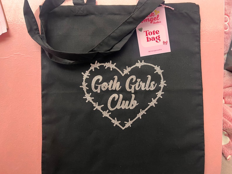 Puede incluir: Bolsa de tela negra con un dise&ntilde;o de coraz&oacute;n con alambre de p&uacute;as a su alrededor y el texto " Goth Girls Club" en purpurina plateada.