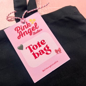 Puede incluir: Bolsa de tela Pink Angel Studios con fondo rosa y texto rojo. El texto dice "Tote Bag" y el nombre de la empresa en las redes sociales es "@pinkangelstudios".