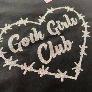 Puede incluir: Una camiseta negra con un dise&ntilde;o de brillo plateado de un coraz&oacute;n de alambre de p&uacute;as que dice "Goth Girls Club" en su interior.