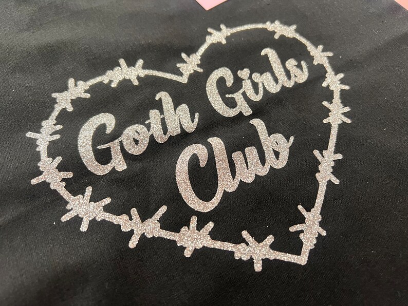 Puede incluir: Una camiseta negra con un dise&ntilde;o de brillo plateado de un coraz&oacute;n de alambre de p&uacute;as que dice "Goth Girls Club" en su interior.