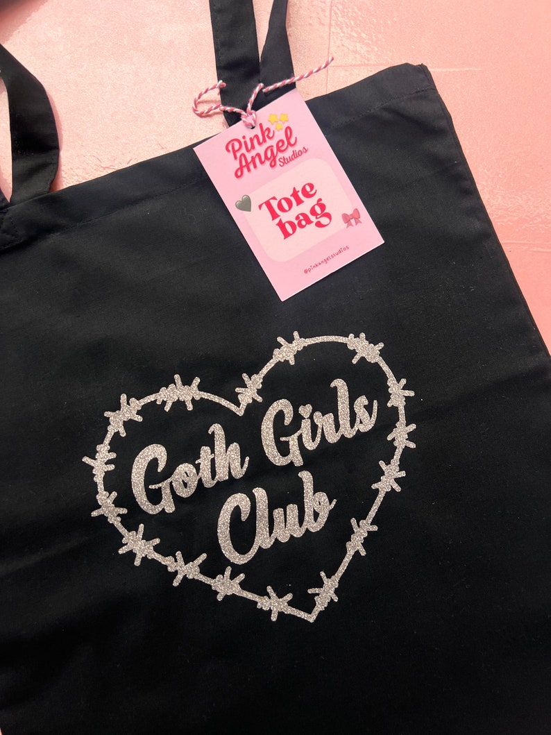 Puede incluir: Bolsa de tela negra con un dise&ntilde;o de brillo plateado de un coraz&oacute;n rodeado de alambre de p&uacute;as y el texto "Goth Girls Club". La bolsa tiene una etiqueta rosa adjunta con el texto "Pink Angel Studios Tote Bag #pinkangelstudios"