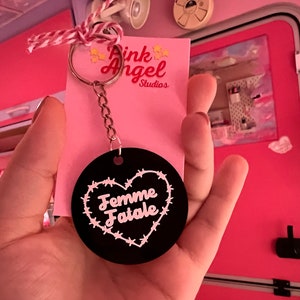 Puede incluir: Un llavero negro y rosa con un dije en forma de corazón que dice "Femme Fatale" rodeado de alambre de púas. El llavero está unido a una cadena plateada y un anillo plateado. El llavero está en una tarjeta rosa con el texto "Pink Angel Studios".