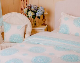 May include: A white bed set with a light blue circular mandala pattern. The set includes a duvet cover, pillowcases, and a decorative pillow on a white wicker chair. A vase of blue and purple flowers sits on a white nightstand.
