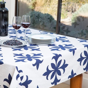 May include: A white tablecloth with a blue floral pattern. Two wine glasses with red wine are on the table, along with a plate of blueberries and a stack of white plates.