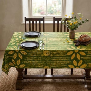 May include: A dining table set for a meal, featuring a green tablecloth with yellow floral patterns. The table is set with dark plates, glasses, and silverware. A vase of flowers and a loaf of bread sit on the table.