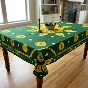 May include: A green rectangular tablecloth with a yellow floral pattern. The tablecloth is on a wooden table with dark brown legs. Two brass candle holders and a bowl of decorative items are on the table.