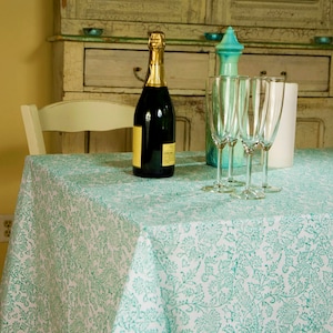 May include: A table setting featuring a bottle of champagne, four champagne flutes, and a decorative turquoise bottle. The table is covered with a white and turquoise patterned tablecloth. The setting suggests a celebratory occasion.