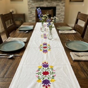 May include: A white table runner with embroidered floral designs in red, yellow, and purple. The runner is placed on a wooden table set with plates, silverware, and napkins. Two vases with flowers are in the center.