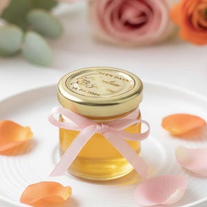 May include: A small glass jar filled with honey, topped with a gold lid and a pink ribbon. The lid has text and a date. The jar sits on a white plate with scattered orange and pink flower petals. Roses are in the background.