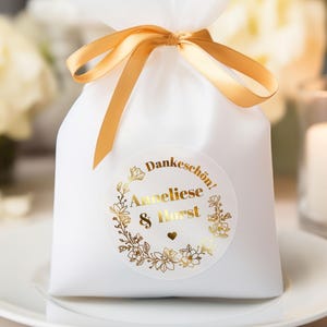 May include: A white gift bag tied with a gold ribbon, featuring a gold foil sticker with the text "Dankesch&ouml;n! Anneliese & Horst" surrounded by floral elements. The bag sits on a white plate, with a lit candle in the background.