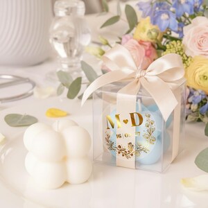 May include: A clear box tied with a satin bow contains light blue candles with gold foil labels. The labels display initials and a date. A white bubble candle sits on a plate, with a bouquet of flowers in the background.