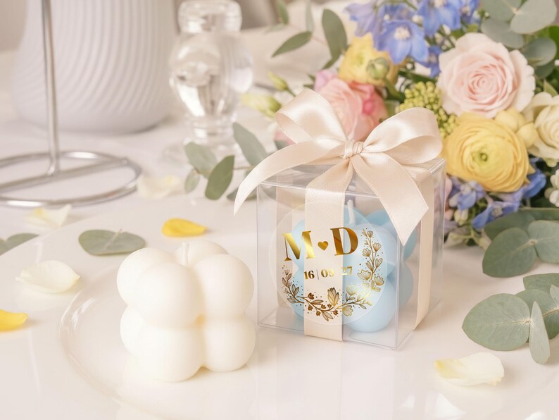 May include: A clear box tied with a satin bow contains light blue candles with gold foil labels. The labels display initials and a date. A white bubble candle sits on a plate, with a bouquet of flowers in the background.