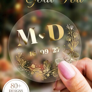 May include: Clear round sticker with gold foil text that reads "Gold Foil". The sticker features the initials "M D" with a heart, date "16 | 09 | 27", and floral design. The sticker also has the text "80+ DESIGNS ZUR AUSWAHL".