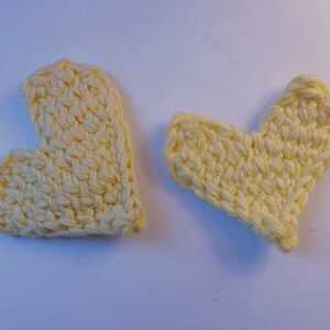 May include: Two handmade, pale yellow crocheted hearts. One heart is upright, and the other is tilted to the right. The hearts are made with a textured stitch, and the yarn appears to be soft. The hearts are likely decorative.