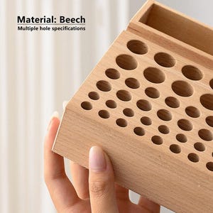 May include: A wooden organiser with multiple holes of different sizes for storing items like paint tubes or makeup brushes. The organiser is made of beech wood and has a small compartment on the top for storing other items. "Material: Beech" and "Multiple hole specifications" are printed on the front of the organiser.