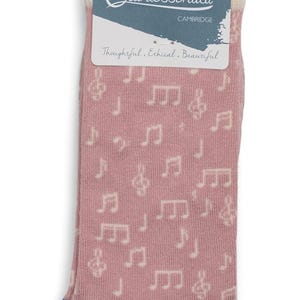 May include: A pair of pink socks with a musical note pattern. The socks have a blue toe and heel, and a cream-colored cuff. The packaging includes the text "The Quintessential Cambridge" and the words "Thoughtful. Ethical. Beautiful."