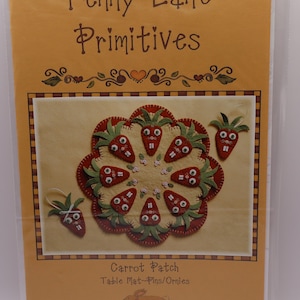 May include: A craft kit titled "Penny Lane Primitives" featuring a carrot-themed design. The kit includes a felt table mat or ornament with carrot faces, in shades of red, green, and beige. The text "Carrot Patch" is visible.