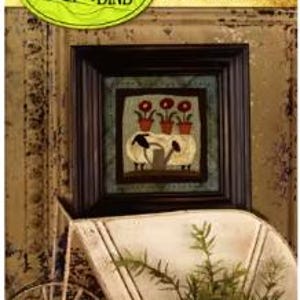 May include: A framed artwork featuring two sheep tending to potted red flowers. The artwork is in a dark wooden frame, displayed on a white wheelbarrow with a small potted plant in front. The artwork is labeled "677 - Wooly Gardener" and "Threads that Bind".