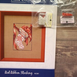 May include: A cross-stitch kit featuring a framed Christmas stocking design with red and white accents. The kit includes beads and the text "Red Ribbon Stocking". The image is on a wooden surface.
