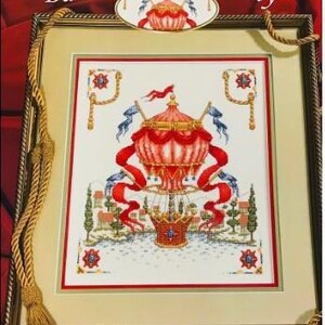 May include: Framed cross-stitch art titled "Balloon Over Tuscany." The artwork features a red and white hot air balloon with blue accents, ribbons, and flags. The scene includes a landscape with trees and buildings, all within a gold frame.