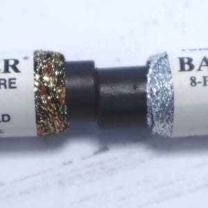 May include: Two spools of Balger 8-Ply Ombre thread. One spool is labeled "Misty Gold" and the other "Silver." The thread is wrapped around a black spool. The labels include the thread's color and the number 1700 and 1000.