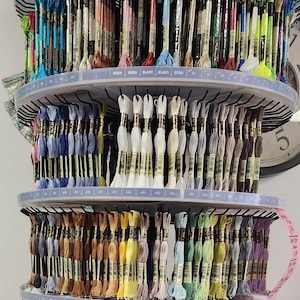 May include: A multi-tiered display of embroidery floss in a variety of colors. The floss is organized by color and number on circular shelves. The colors range from blues and greens to yellows and browns. The display is well-lit and organized.