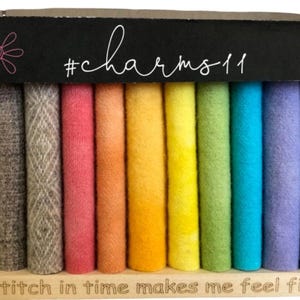 May include: A rainbow assortment of felt rolls in a wooden display. The top of the display has the text "#charms11" and a floral design. The bottom of the display has the text "A stitch in time makes me feel fine."