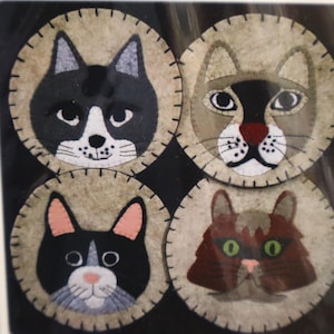 May include: Four round cat-themed coasters. Each coaster features a different cat face design, with varying colors including black, brown, and tan. The coasters have a stitched border and are set against a dark background.