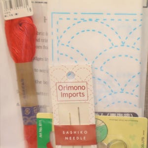 May include: A collection of sewing supplies, including a spool of red thread, a package of Sashiko needles, a Clover thimble, and a white fabric with a blue dotted design. The items are packaged in clear plastic.