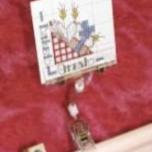 May include: A cross-stitch project featuring the word "Fresh" and a design of wheat and a checkered pattern. The project is held in place by a wooden frame and clips, set against a textured red background.