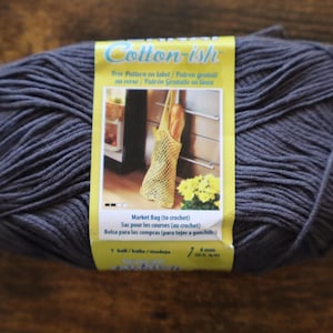 May include: A large ball of dark gray Cotton-ish yarn with a yellow label. The label features text in multiple languages and an image of a crocheted market bag holding a baguette. The yarn is 4 mm thick.