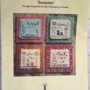 May include: A cross-stitch pattern titled 'Seasons' with designs for Spring, Summer, Autumn, and Winter. Each season is represented by a square with embroidered elements like flowers, animals, and seasonal text. The Haystack brand is visible.