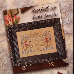 May include: A framed cross-stitch sampler titled "Rosa Smith 1840 Basket Sampler." The sampler features a detailed floral design with blue, red, and tan hues. The frame is dark brown with ornate detailing, and the background is a floral pattern.