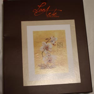 May include: A boxed cross-stitch kit featuring a floral design. The artwork showcases white flowers with a gold background, framed in a white mat. The box is brown with the text "Lanarte the Art of Stitching" in orange.