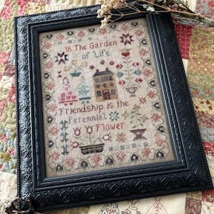 May include: Framed cross-stitch artwork with the text "In The Garden of Life" and "Friendship is the Perennial Flower." The artwork features a house, flowers, and figures in shades of red, blue, and green. The frame is black.