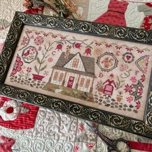 May include: Framed cross-stitch sampler featuring a house, flowers, and birds in a detailed, folk art style. The sampler is in a dark green ornate frame. The background is a quilted patchwork with red and white patterns. A pair of scissors rests on the quilt.