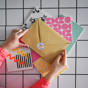 Può includere: Un mucchio di carta con motivi colorati con una busta marrone sopra. La busta ha un adesivo a forma di cuore sul davanti.