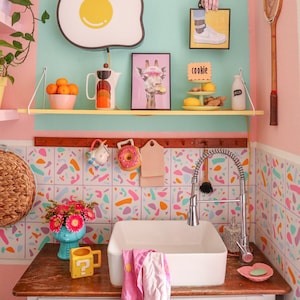 Könnte beinhalten: Ein weißes Küchenspülbecken mit einem verchromten Wasserhahn und einer rosafarbenen und weißen Fliesenrückwand. Das Spülbecken befindet sich in einer Küche mit einem rosa-blauen Farbschema. Auf den Regalen über dem Spülbecken befinden sich verschiedene Dekorationsartikel, darunter ein gelber, eiförmiger Teller, eine Giraffenfigur und eine Keksdose.