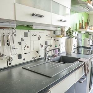May include: A black kitchen sink with a chrome faucet, a black countertop, and white cabinets. The backsplash is white tile with black geometric shapes.