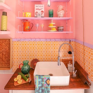 Könnte beinhalten: Ein weißes Porzellanwaschbecken mit einem verchromten Wasserhahn und einer Holztheke. Das Waschbecken ist an einer rosa Wand mit einem Regal darüber angebracht. Das Regal ist mit verschiedenen Gegenständen gefüllt, darunter eine rot-weiße Schachtel mit dem Text "Mutter, hast du Kellogg's gesagt?" und eine grüne Glaskaraffe. Die Wand hinter dem Waschbecken ist mit einem Muster aus orangen Kreisen und violetten Quadraten gefliest.