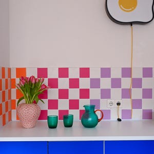 Op de afbeelding: Een keukeninterieur met een blauwe onderkast, een wit aanrechtblad en een kleurrijke tegelachterwand. Een roze vaas met roze tulpen, twee groene glazen en een turquoise kan met een oranje handvat staan op het aanrecht. Een eivormige lamp hangt erboven.