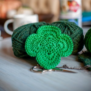 May include: A green crocheted clover charm with two balls of green yarn and a pair of silver scissors on a white wooden surface.